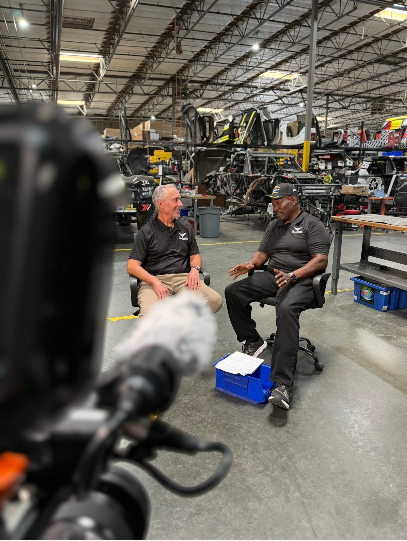Camera and microphone facing two seated interview subjects inside a workshop